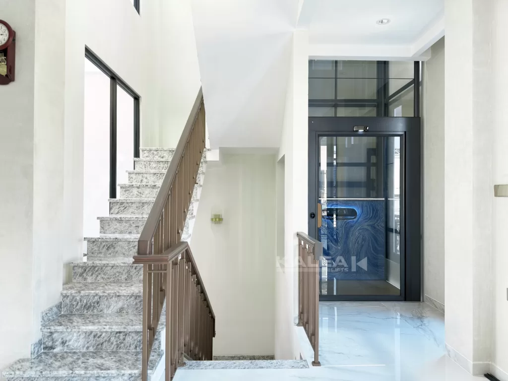 Lift rumah Kalea dengan shaft kaca hitam dipasang di samping tangga marmer pada area void rumah modern bertingkat