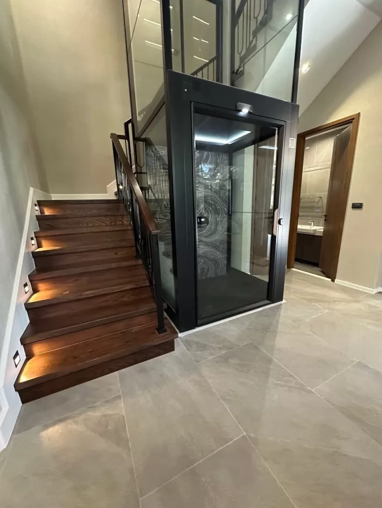 Kalea X80 Umbra sebagai salah satu jenis lift rumah dengan desain kabin elegan, ditempatkan di samping tangga untuk hunian modern yang membutuhkan akses vertikal yang nyaman